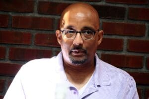 A man with glasses and bald head in front of a brick wall.